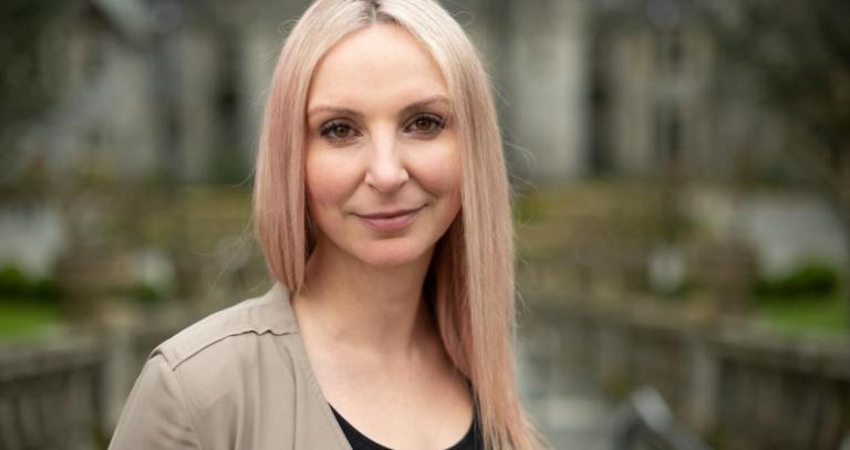 A woman with a small smile looks at the camera. A stone building is in the background.