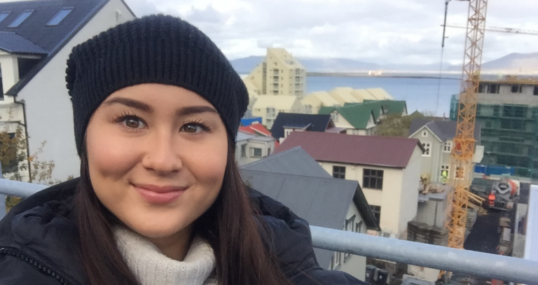 A woman in a toque smiles from what might be a balcony. Homes and a crane can be seen in the distance below.