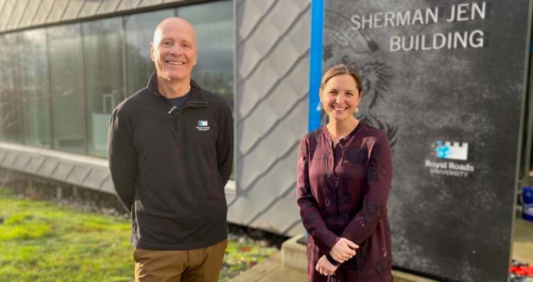 A man and a woman pose smiling in front of the entrance to a building.