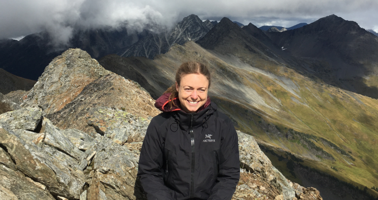 Kristina Schrange poses on a rocky peak wearing a black jacket. High altitude clouds and a green mountain slope are in the background. 