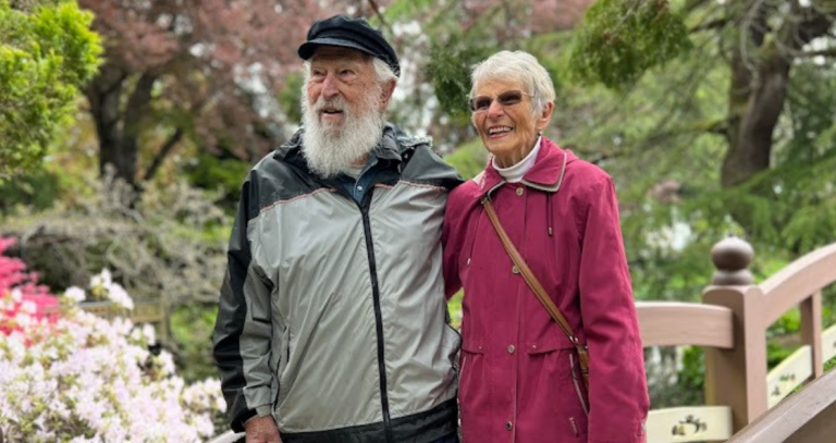 Dick (left) and Bette (right) Geisreiter standing arm in arm on a bridge in the Royal Roads Japanese Garden.