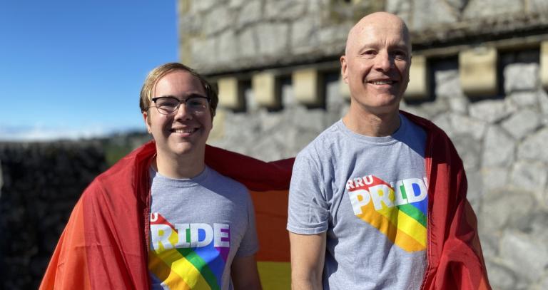 President Steenkamp and student Shaun Fisk draped in the Progress Pride flag atop Hatley Castle. Both are wearing RRU Pride t-shirts and the skies are blue..