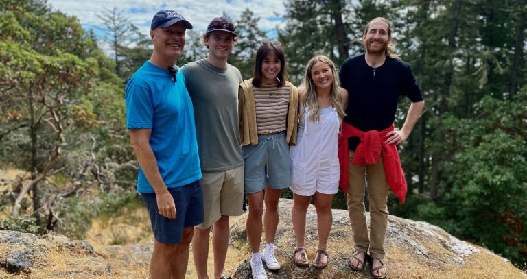 President Philip Steenkamp with four students atop a Garry Oak meadow that looks over the trees and Esquimalt Lagoon.