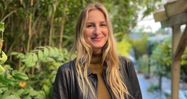 Danica Straith stands in front of a natural background with greenery and a path. She's wearing a brown sweater and a black leather jacket. 