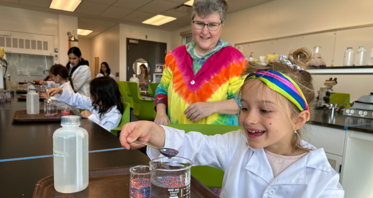 Mickie Noble in tie dye lab smock standing beside young girl mixing hydrophic sand and water.