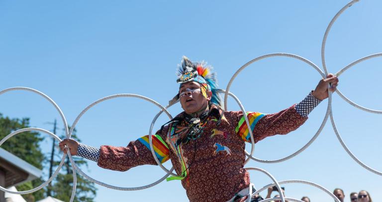 An Indigenous hoop dancer performing at an outdoor event under a clear blue sky. The dancer is wearing a traditional outfit with vibrant colors and intricate patterns, including a headdress with feathers and a decorated shirt. 