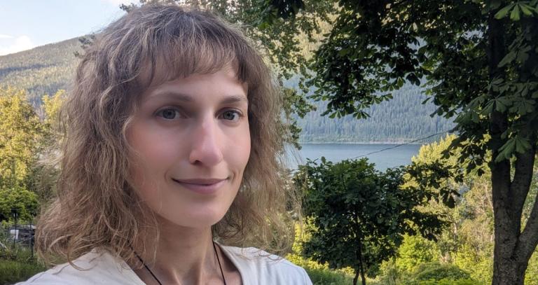 Margot Webster stands close to the camera; framed behind her is a beautiful nature scene with abundant green trees and the glistening blue of a lake in the background