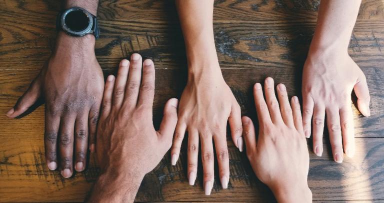 Aerial image of five hands in different skin tones laying palm down on a wooden surface.