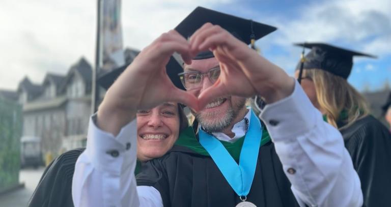 Two graduates smile while peeking through heart hands.