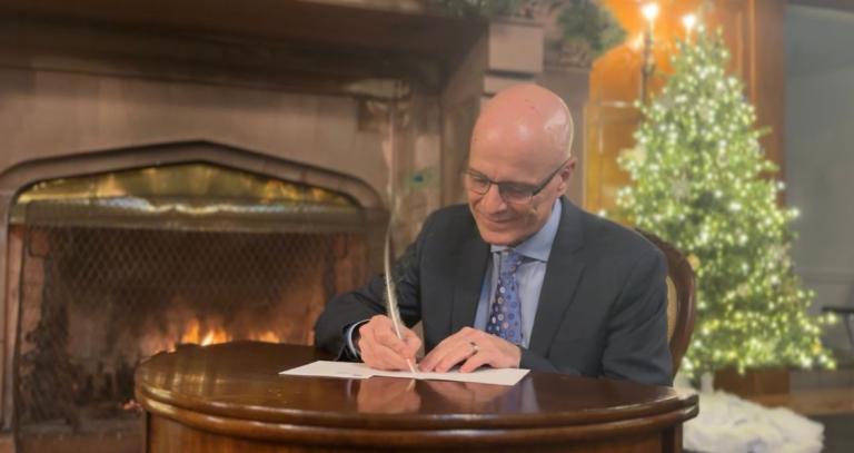 A man is seen writing at a desk with a peacock feather.