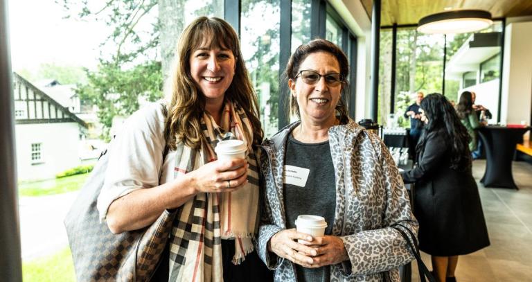 Two alumni stand in front of the windows in the Dogwood Building, holding disposable coffee cups