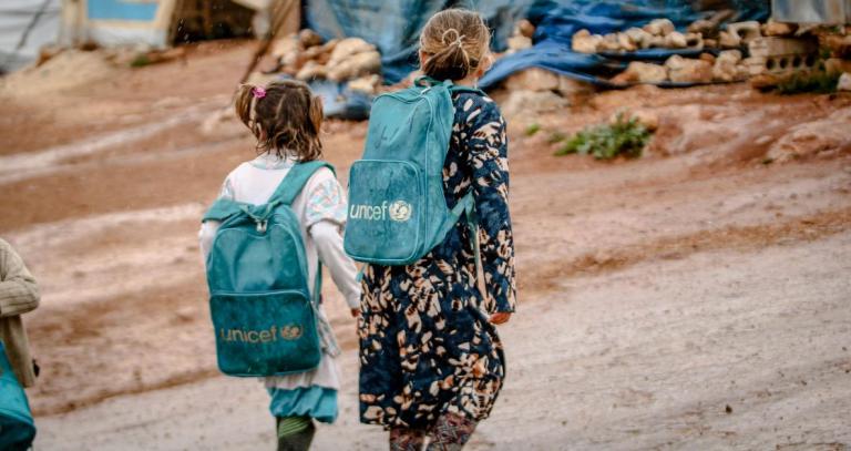 Two children walk through an encampment wearing Unicef backpacks