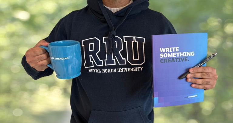 A photo of a man from the neck down holds a notebook in one hand and a mug in the other.