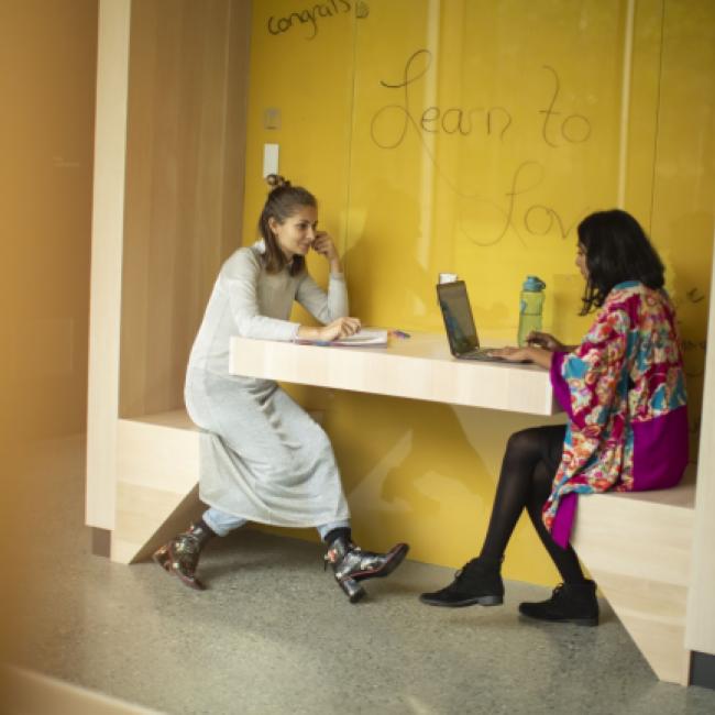 Two students sit in a booth with a yellow background in the Sherman Jen Building.