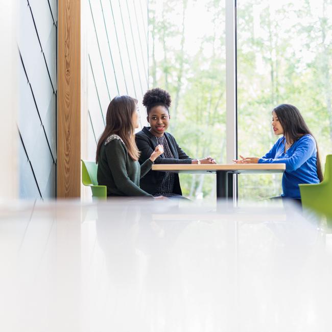 Three-students-sitting-talking-next-to-window-and-forest