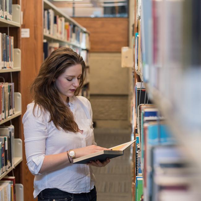 Student holding open book standing in library stacks