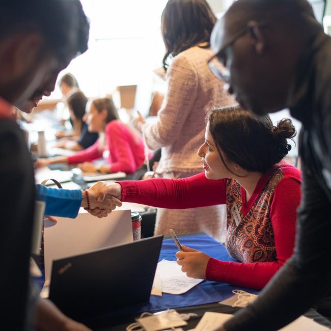 Two people shake hands over a desk with computers while others look over.