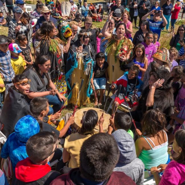 A packed group of singing people in many bright colours surround drum players singing and cheering.