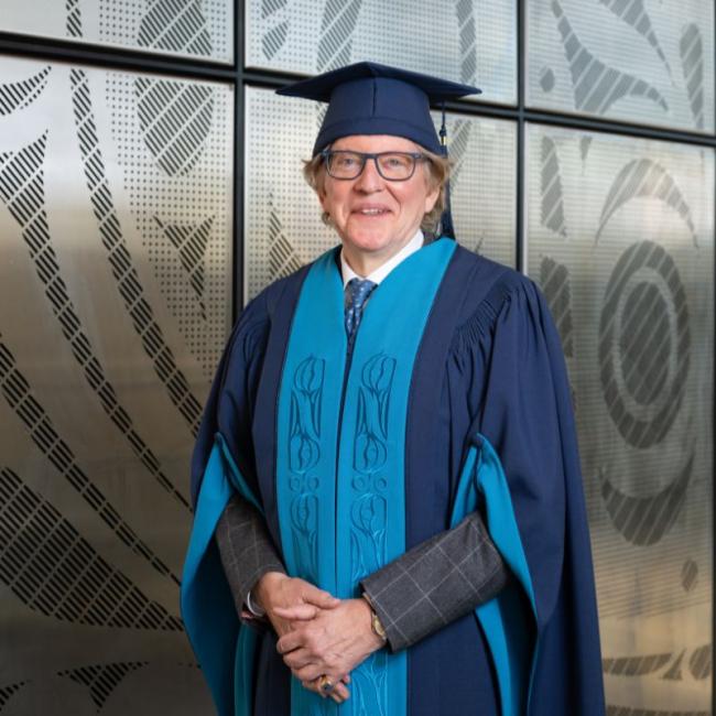 Anthony von Mandl smiling in front of "All My Relations" installation wearing a cap and gown.