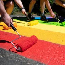 Several hands holding paint rollers, painting a rainbow crosswalk being painted at RRU.