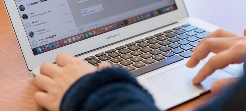 A silver laptop, opened to a social media page, is viewed from over someone's shoulder. The person's hands and black sleeves are visible over the keyboard.